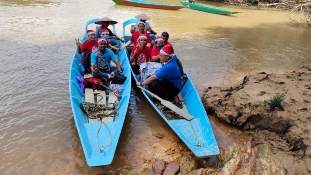 Two blue longboats in Malaysia. In them sit a missionary Priest and several volunteers from his Parish, all wearing santa hats. they are preparing to travel up the river to bring practical help and Christmas hope to remote communities